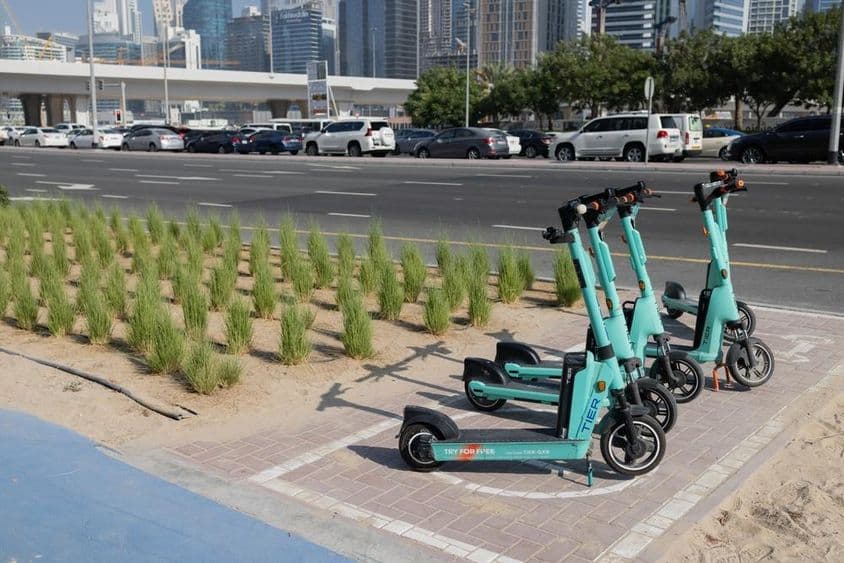 E-scooters estacionados em frente a arranha-céu de Dubai com plantas verdes no separador de pista.