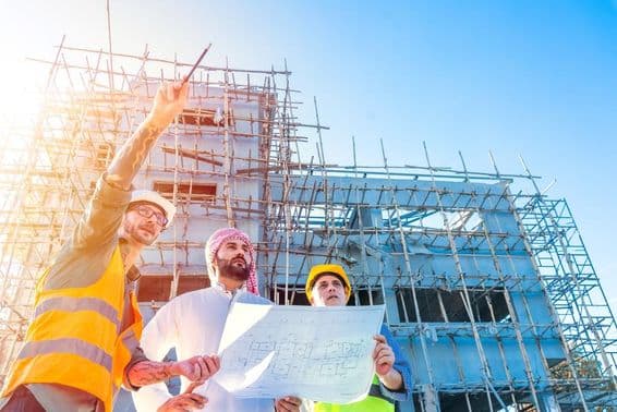 Construção de casa em Dubai, edifício com andaimes em frente, cidadão Emirati segurando uma planta, consultando com dois engenheiros de construção.