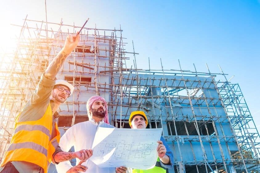 Construção de casa em Dubai, edifício com andaimes em frente, cidadão Emirati segurando uma planta, consultando com dois engenheiros de construção.