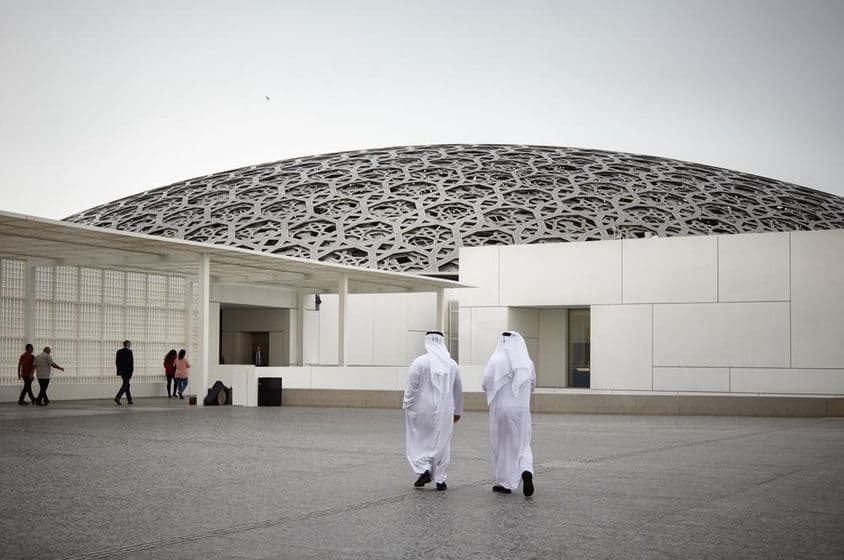Turistas no museu Louvre de Abu Dhabi e duas pessoas com trajes tradicionais Emiradenses brancos.