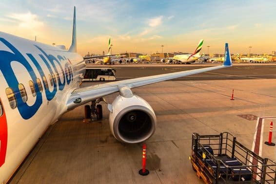 Avião da Flydubai estacionado com diversos outros aviões ao fundo e um ônibus de transporte de passageiros, à direita com bagagens em um pequeno veículo de transporte.