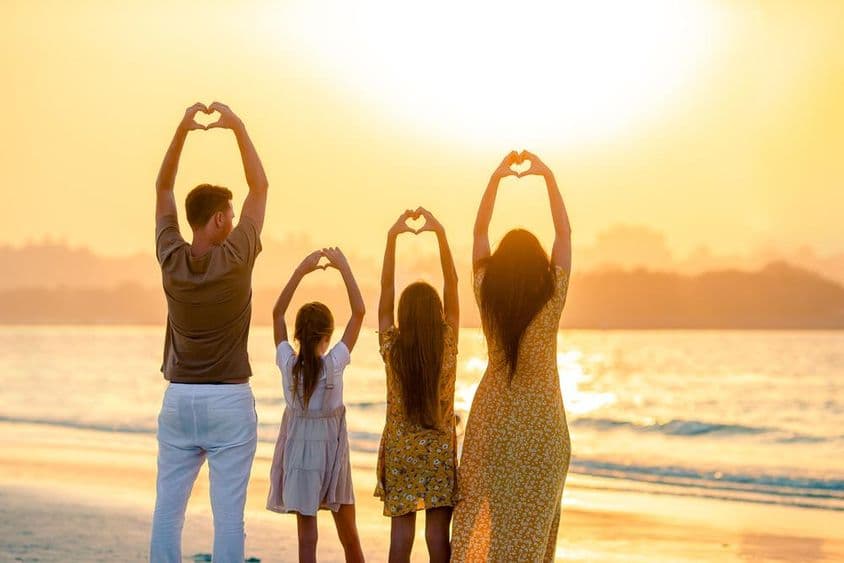 Pessoas na praia, de costas para a câmera, formando um coração com as mãos, com o mar ao fundo.