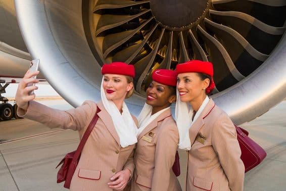 Três comissários de bordo da Emirates tirando uma selfie em frente a um motor de avião da Emirates.