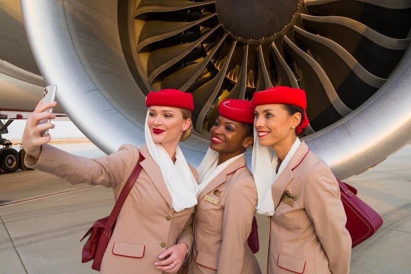 Três comissários de bordo da Emirates tirando uma selfie em frente a um motor de avião da Emirates.