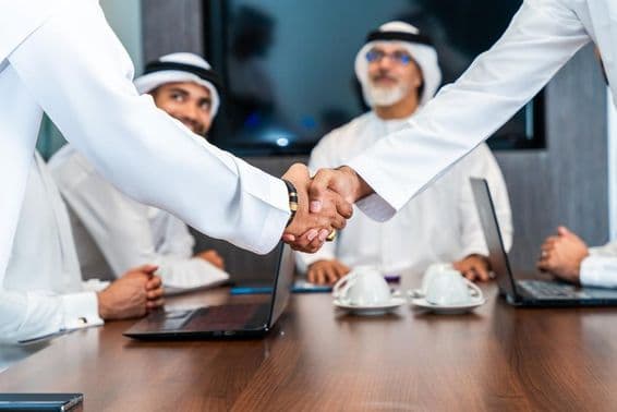 Executivos emiradenses apertam as mãos em uma mesa de conferência, com telefones, cadernos e xícaras de café sobre a mesa. Dois emiradenses sorriem ao fundo.