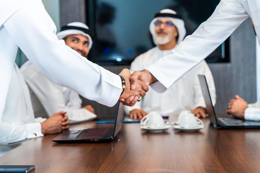 Executivos emiradenses apertam as mãos em uma mesa de conferência, com telefones, cadernos e xícaras de café sobre a mesa. Dois emiradenses sorriem ao fundo.