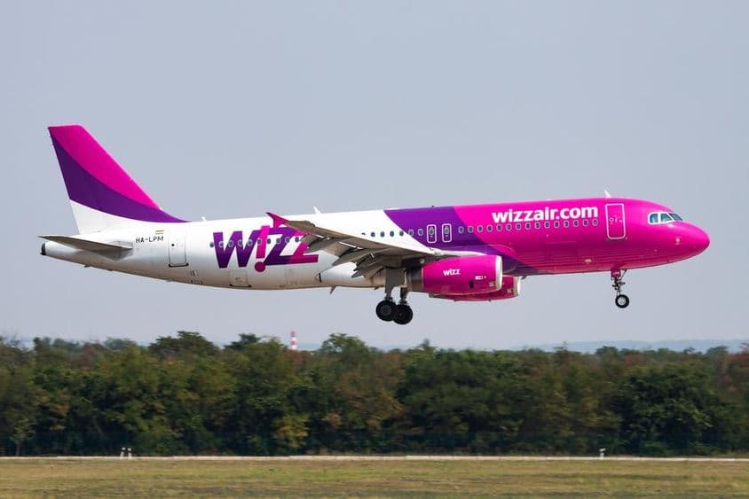 Avião da Wizz Air voando em baixa altitude com floresta ao fundo.