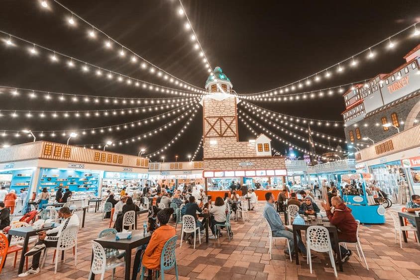 Global Village, turistas e locais degustando especialidades gastronômicas em mesas.