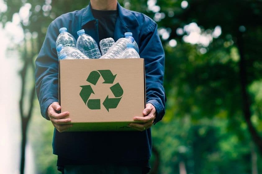 Uma pessoa carregando uma caixa de papelão com garrafas retornáveis, com árvores verdes desfocadas ao fundo.