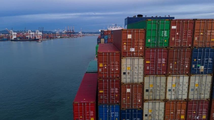 À esquerda, uma baía de um porto marítimo é visível, à direita contêineres empilhados.