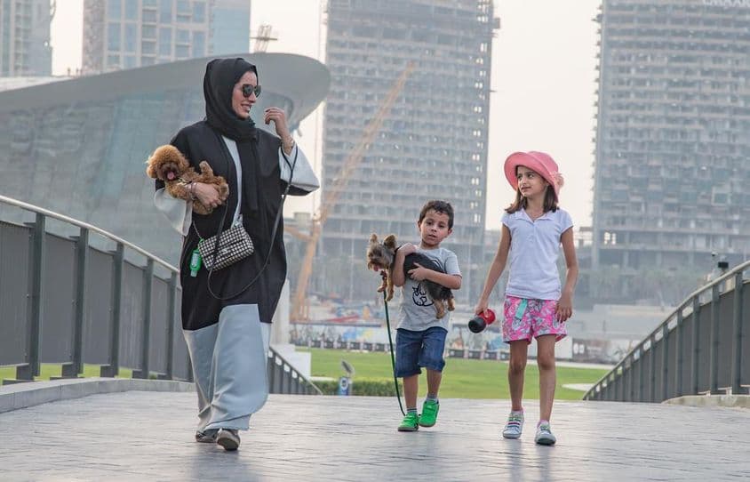 Uma família de quatro pessoas em frente aos arranha-céus de Dubai, com a mãe e uma criança segurando um cachorro.