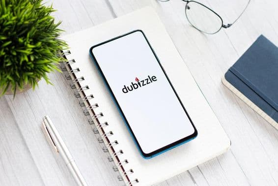 Um telefone com Dubizzle na tela sobre uma mesa, sob um bloco de notas, próximo a óculos e uma pequena planta verde.