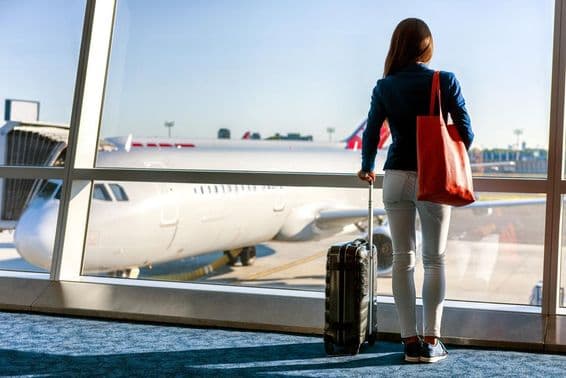 Uma mulher está de pé junto à janela com um avião visível, segurando uma mala de rodinhas com a mão esquerda e uma bolsa na direita.
