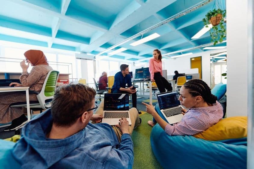 Pessoas trabalhando em um escritório, duas pessoas sentadas em puffs com laptops no colo.