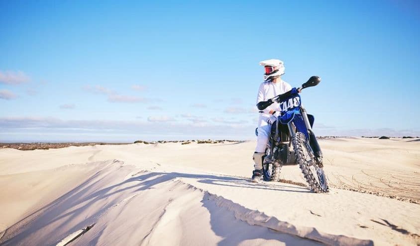 Pessoa pilotando moto no deserto, vestindo roupas brancas em uma motocicleta azul.