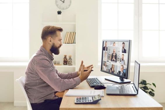 Uma pessoa em um ambiente de escritório com notebook e calculadora na mesa, conduzindo uma videoconferência em um monitor com webcam.