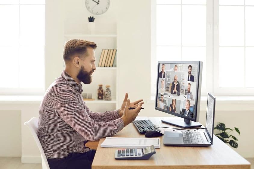 Uma pessoa em um ambiente de escritório com notebook e calculadora na mesa, conduzindo uma videoconferência em um monitor com webcam.