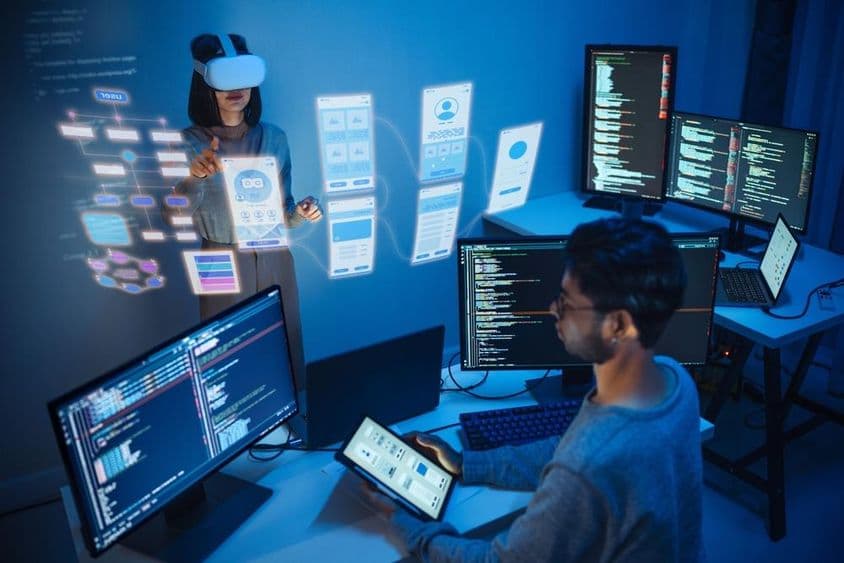 Um desenvolvedor de aplicativos sentado em frente a um computador, segurando um tablet, com dois monitores exibindo código de computador.