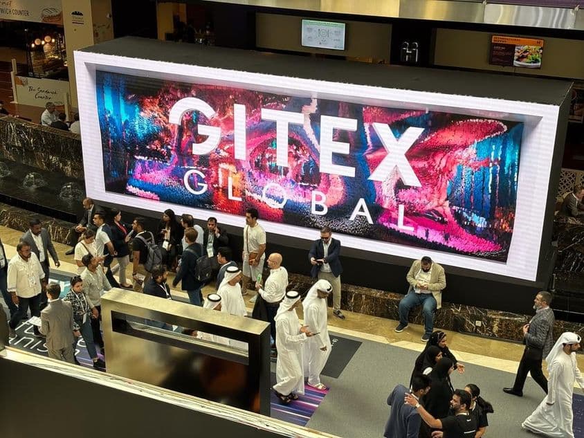 Gitex Global ao fundo com pessoas, principalmente homens e mulheres Emiratis em primeiro plano.
