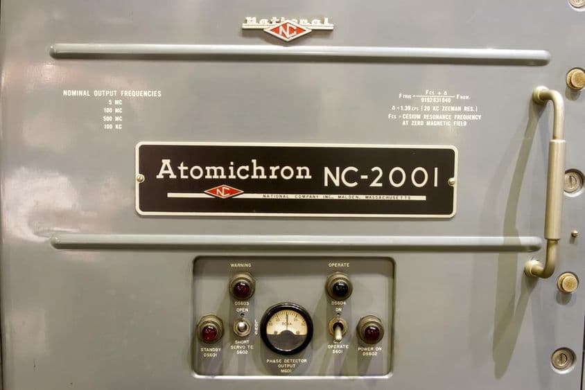 Exibido está o painel de controle de um antigo relógio atômico, junto com seu nome em uma pequena placa.