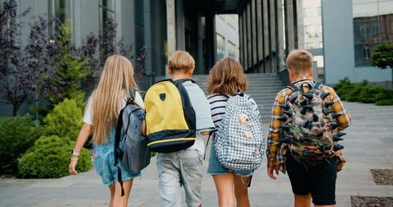 Estudantes carregando mochilas em frente à entrada de uma escola.