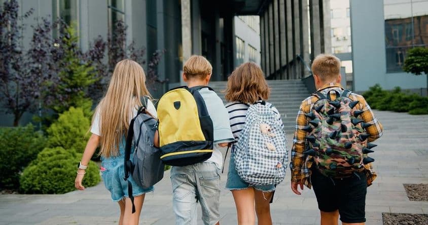 Estudantes carregando mochilas em frente à entrada de uma escola.