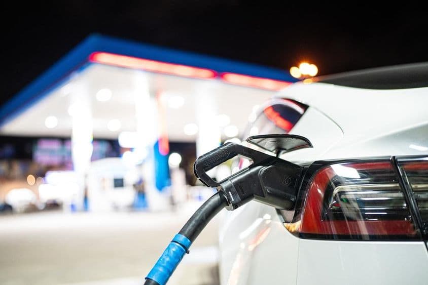 Um Tesla sendo carregado, com um posto de gasolina ao fundo.