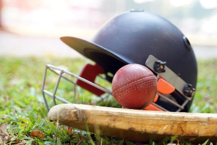 Um taco de críquete com uma bola acima e atrás dele, um capacete de críquete visível em um campo gramado.