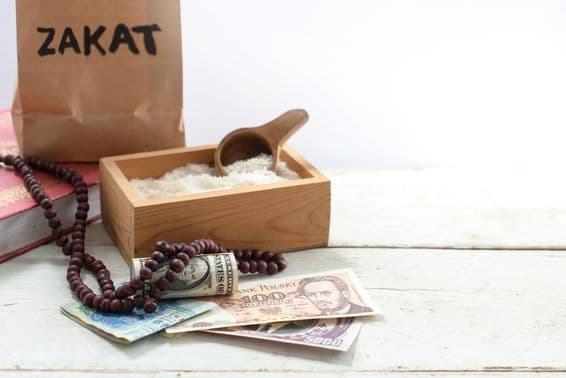Bolsa rotulada como Zakat na frente de uma caixa com arroz, contas de pérolas, dólares e outras moedas.