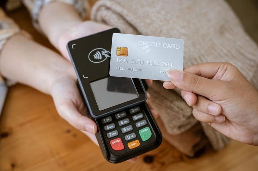 Um cartão bancário segurado por uma mão, com um leitor de cartão ao fundo.