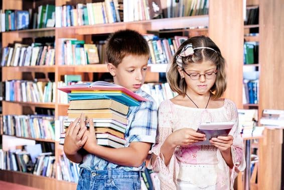 Duas crianças na biblioteca, uma lendo um e-book, a outra carregando muitos livros nas mãos.