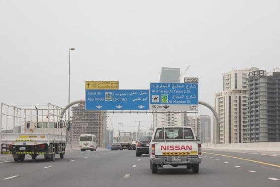 Placa de sinalização rodoviária de Al Khail, com alguns prédios altos ao fundo.