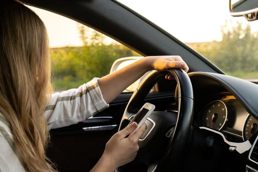 Uma mulher usando um celular enquanto dirige, segurando o volante com outra mão.