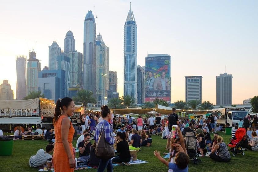 Pessoas em Dubai sentadas em um parque, arranha-céus ao fundo.