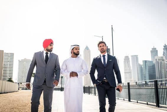 Um indiano, um emirati e um cidadão europeu caminhando ao longo do canal, arranha-céus ao fundo.