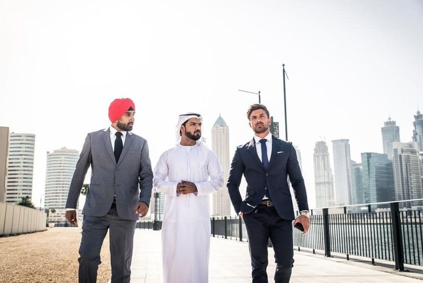 Um indiano, um emirati e um cidadão europeu caminhando ao longo do canal, arranha-céus ao fundo.