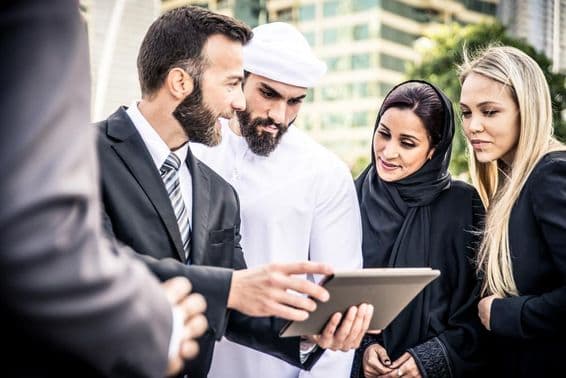 Um homem europeu e um emiradense olhando para um tablet, flanqueados por duas mulheres, uma loira e uma emiradense, observando.