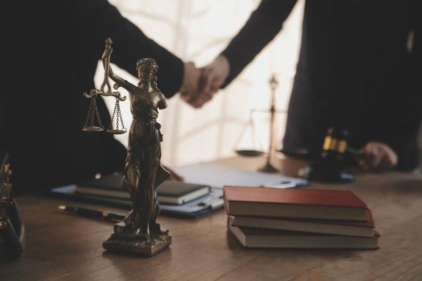 Um advogado é retratado negociando com um cliente, com uma estátua de Justitia visível na mesa.
