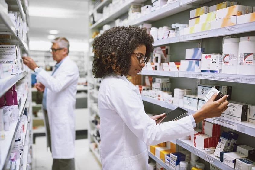 Uma prateleira de farmácia sendo abastecida por uma mulher e um homem verificando medicamentos.