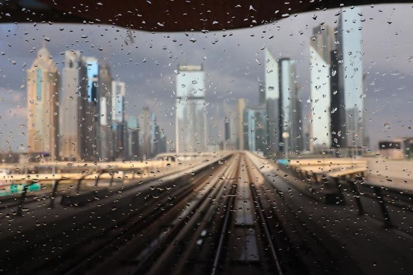 Chuva vista através de uma janela do metrô de Dubai com arranha-céus ao fundo.