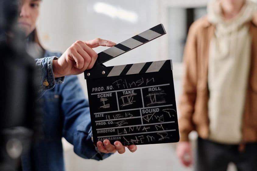 Claquete de filme com escrita de giz, duas pessoas ao lado dela.
