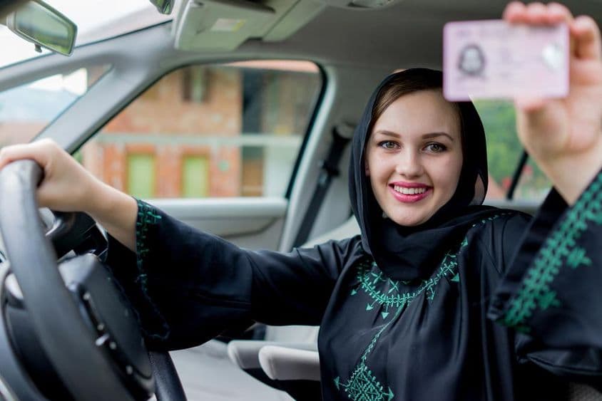 Uma mulher em Dubai segura uma carteira de motorista com uma mão e o volante com a outra, sorrindo.