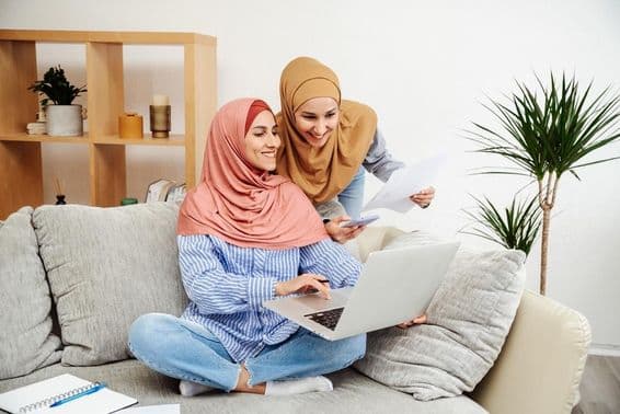 Duas mulheres Emirati revisando um contrato e caderno, uma segurando um telefone, sentadas em um sofá.