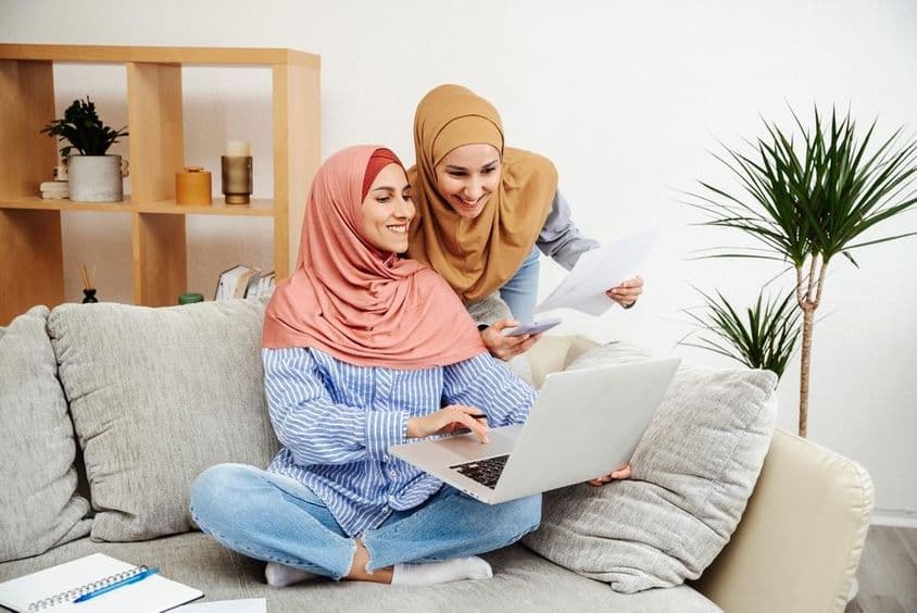 Duas mulheres Emirati revisando um contrato e caderno, uma segurando um telefone, sentadas em um sofá.