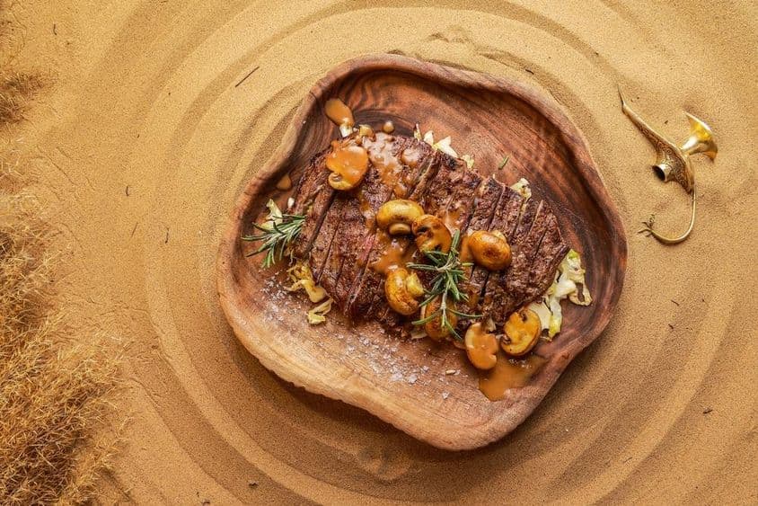Filé servido com cogumelos em uma tábua de madeira na areia do deserto, com uma lanterna apagada ao lado.
