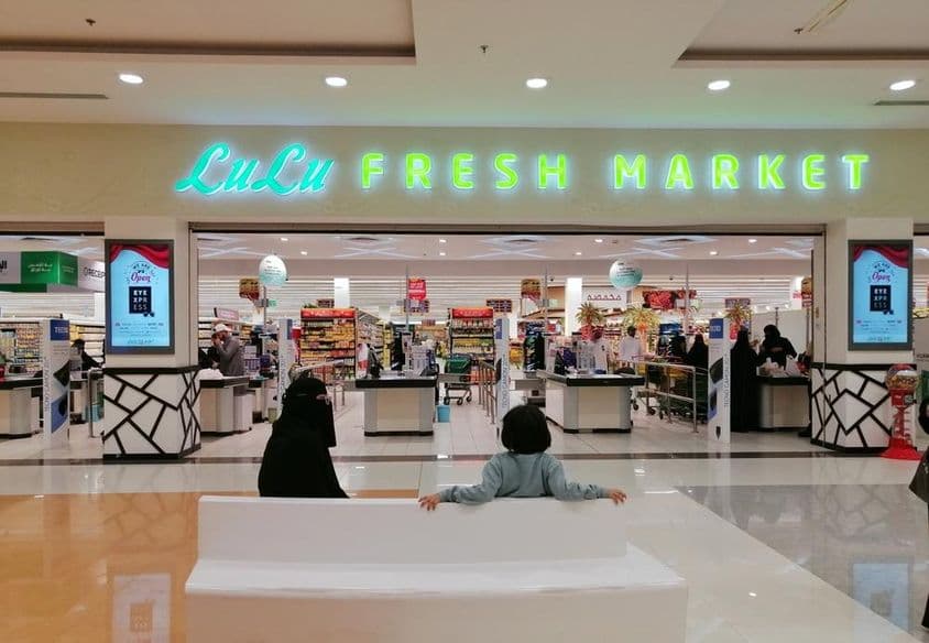 Entrada de uma loja LuLu em um shopping, com duas pessoas sentadas, uma mulher e uma criança, vistas de costas.