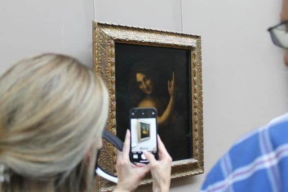 A pintura de São João Batista na parede, com uma mulher tirando uma foto dela com um celular.