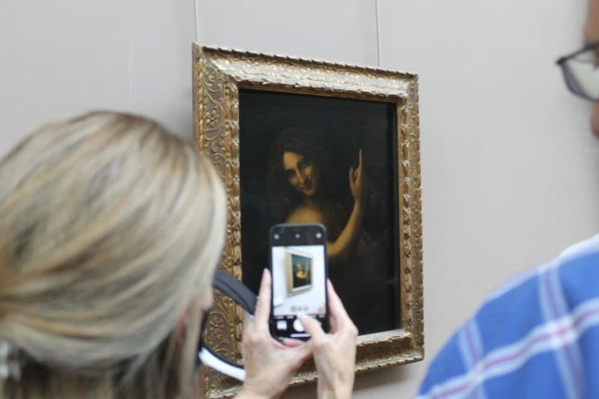A pintura de São João Batista na parede, com uma mulher tirando uma foto dela com um celular.