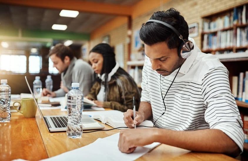 Estudantes universitários escrevendo em papel com livro e caderno à frente, um usando fones de ouvido.