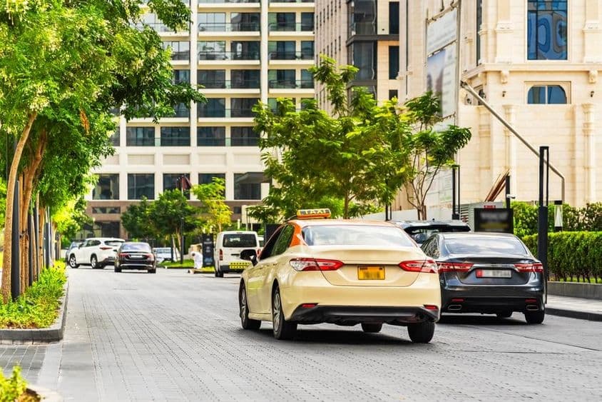 Carros em Dubai na estrada, um táxi em primeiro plano, casas ao fundo.
